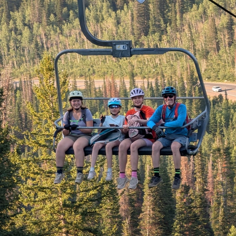 Four people in helmets ride a chairlift over a forest with a road visible in the distance.