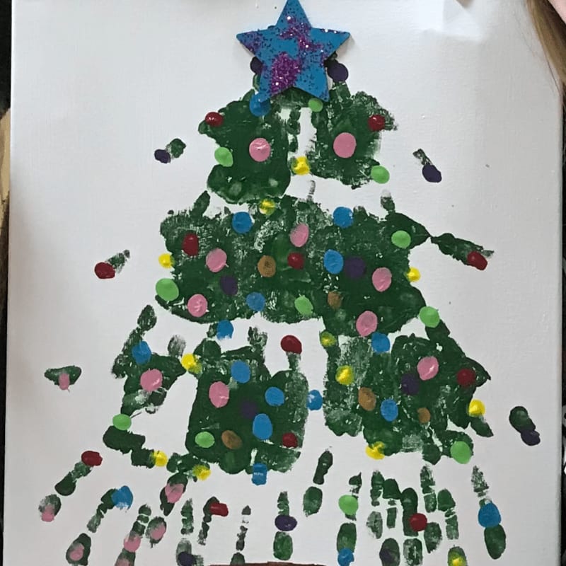 A Christmas tree made of green handprints, adorned with colorful fingerprint ornaments and a glittery star, stands against a white background.