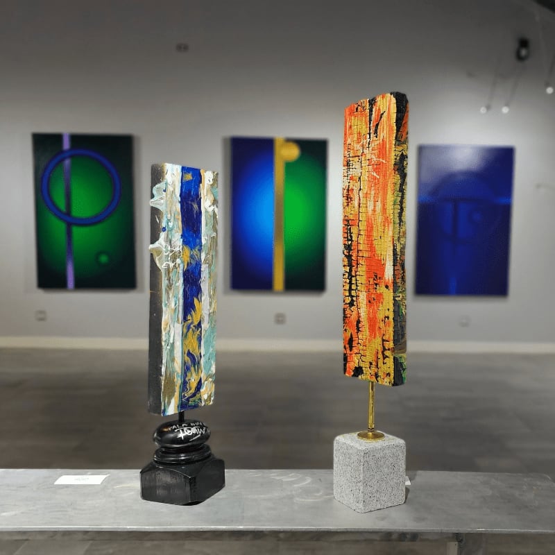 Two abstract sculptures stand on a shelf in the foreground, with a gallery wall displaying colorful abstract paintings in the background.