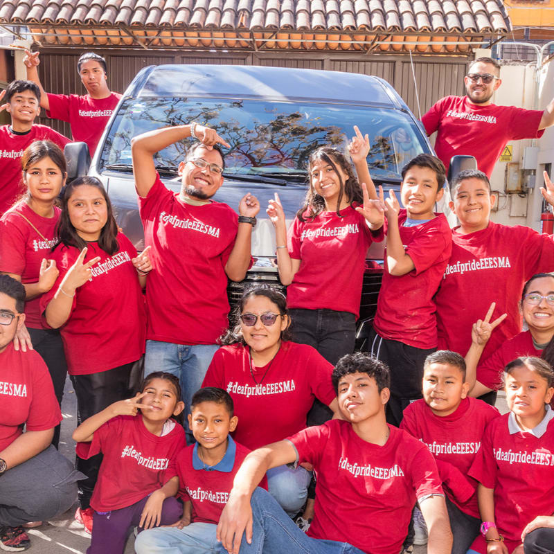 A group of smiling people in red t-shirts pose in front of a van, with a building visible in the background.