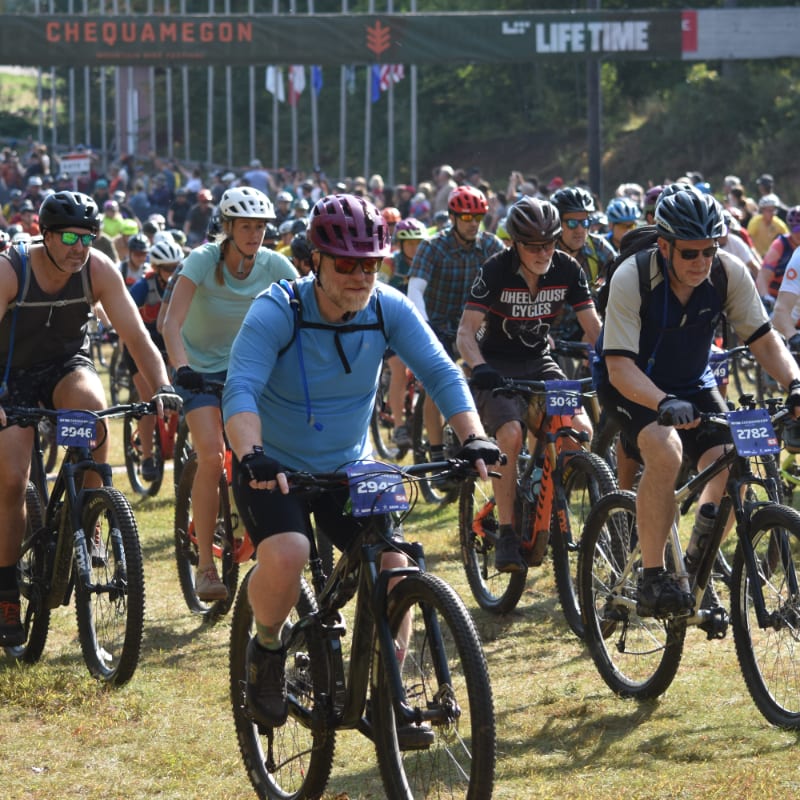 Cyclists in the foreground ride mountain bikes towards a finish line with spectators and flags in the background.