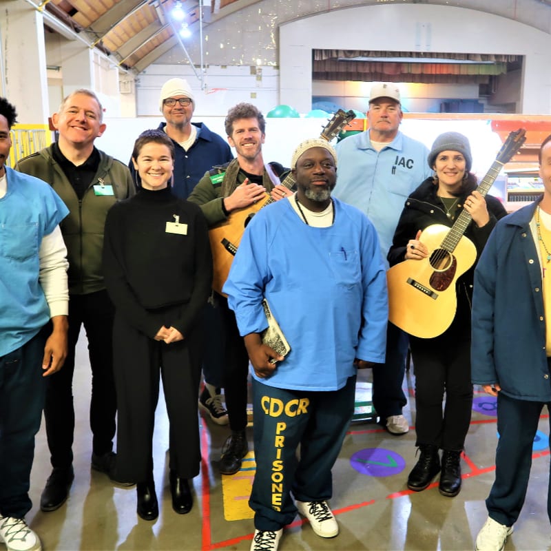 A group of people, some in prison uniforms and others in casual attire, stand together in a large room with musical instruments, while a colorful floor pattern is visible in the foreground and a blurred background suggests an indoor space.