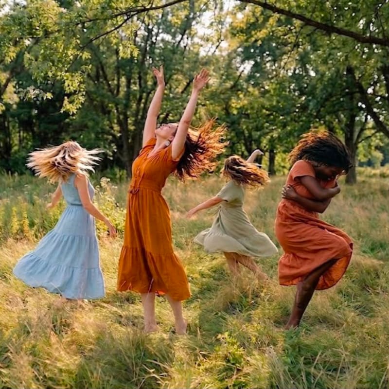 Four women in flowing dresses dance joyfully in a sun-drenched meadow with a lush green forest in the background.