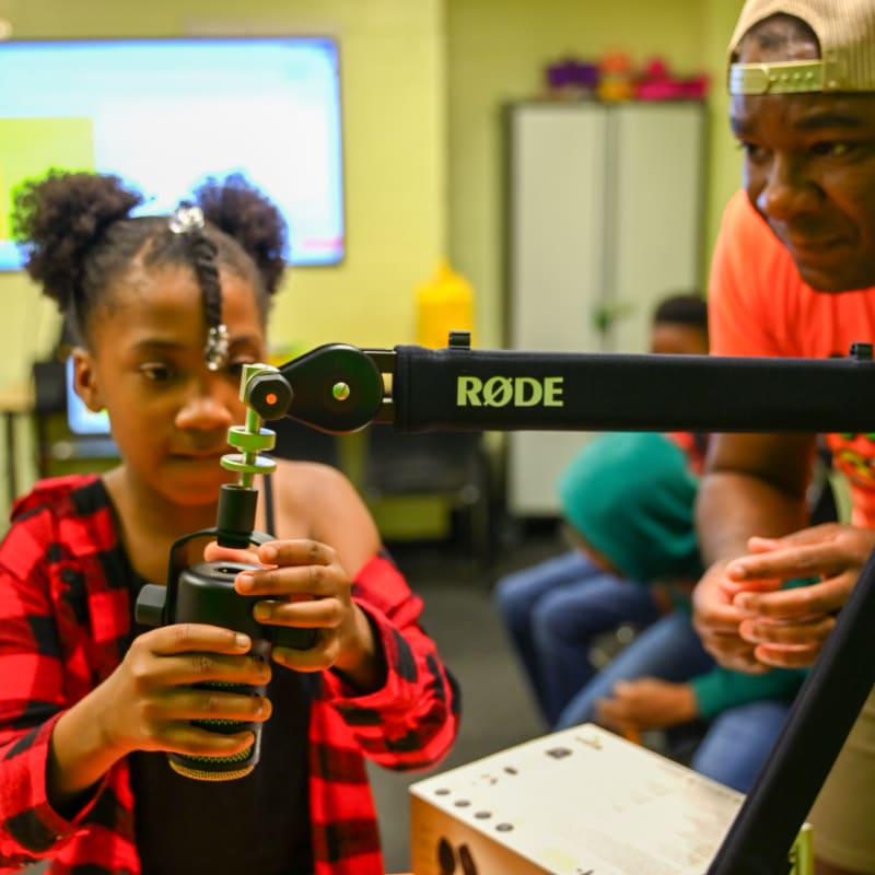 A young girl in a red and black plaid shirt adjusts a microphone in the foreground, while a man in an orange shirt and a blurred background of children and a classroom setting are visible.
