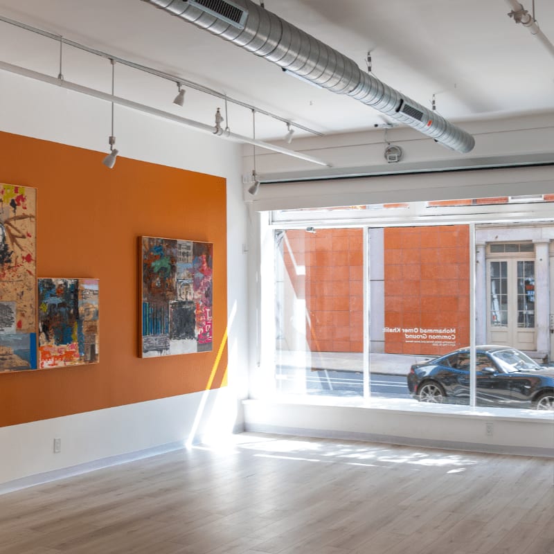 Sunlight streams across the wooden floor of an art gallery, illuminating abstract paintings on an orange wall, while a car passes by the large window in the background.