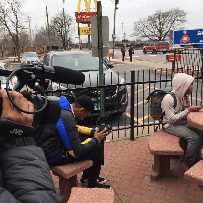 In the foreground, a person films with a professional camera, while in the background, two young men sit at a picnic table outside a McDonald's.