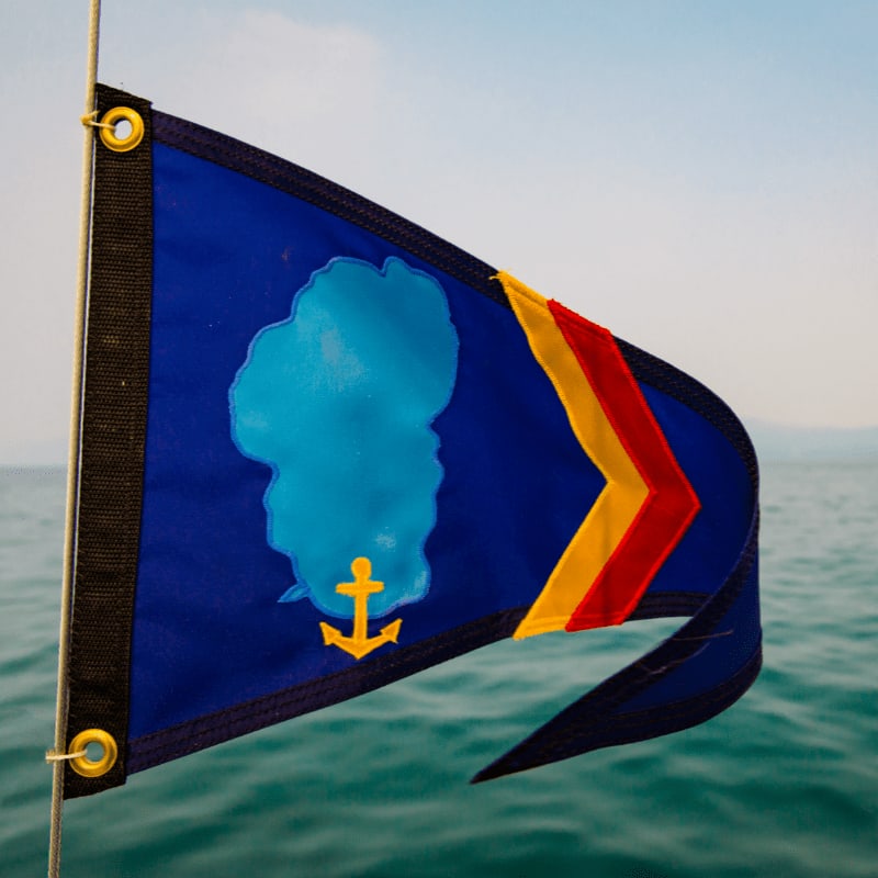 A blue triangular flag with a light blue silhouette and a yellow anchor is fluttering in the foreground against a backdrop of calm blue water and a hazy sky.
