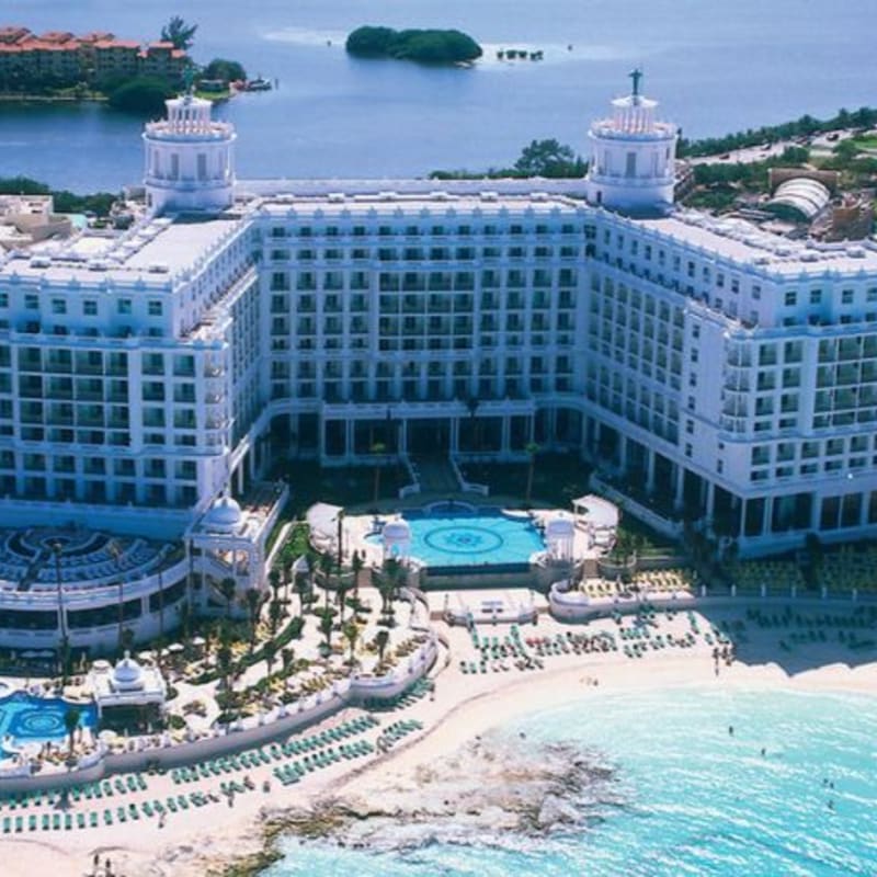 A grand white hotel with a pool and beach in the foreground overlooks a tranquil blue bay in the background.