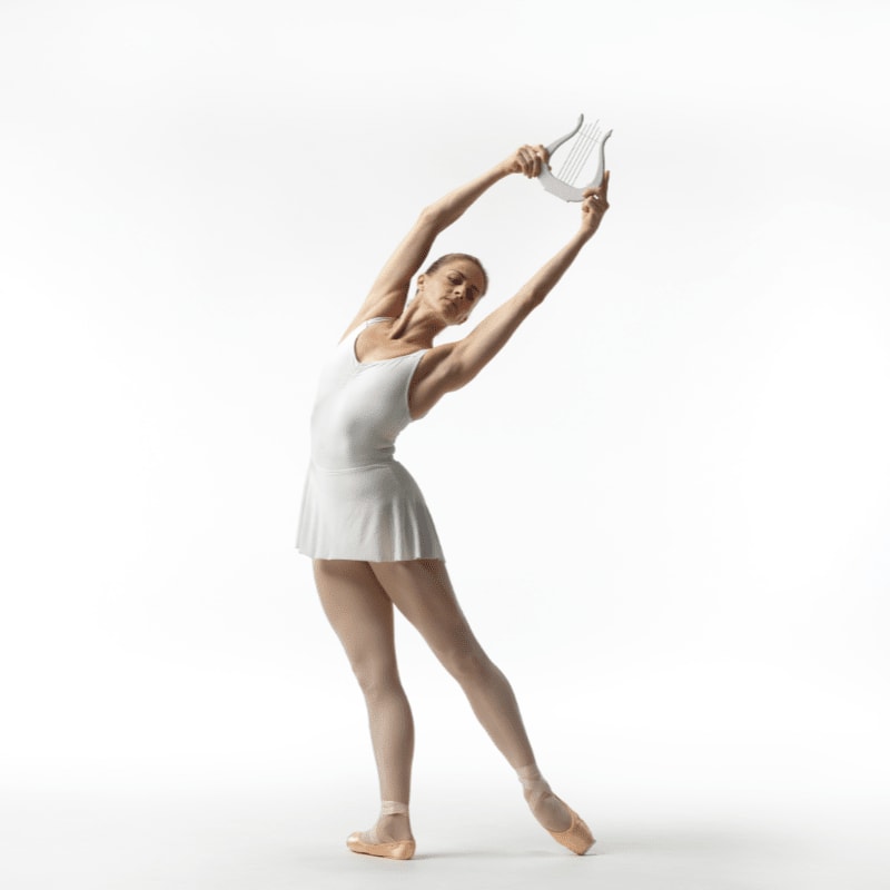 A ballerina in a white leotard and skirt gracefully poses with a lyre held above her head against a plain white background.
