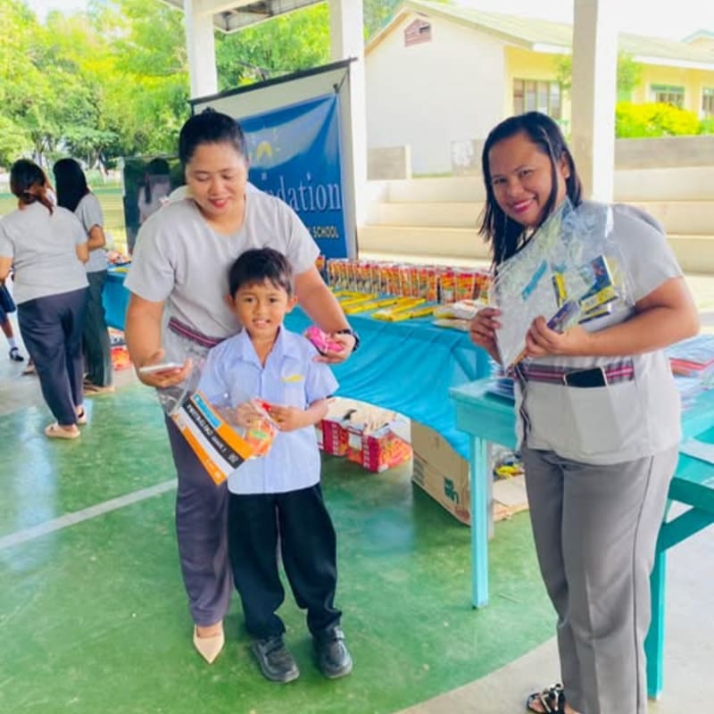 Two women and a young boy stand in the foreground holding school supplies, with a table laden with more supplies and a banner in the background.
