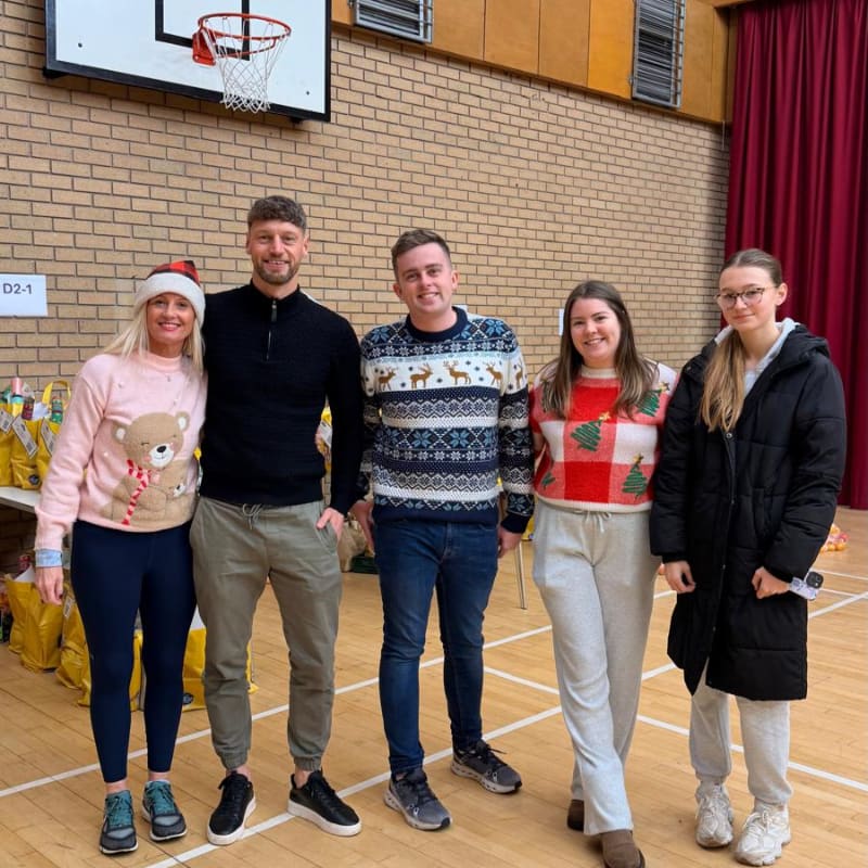 Four people in festive sweaters stand in a gymnasium with a basketball hoop above and gift bags to their left.