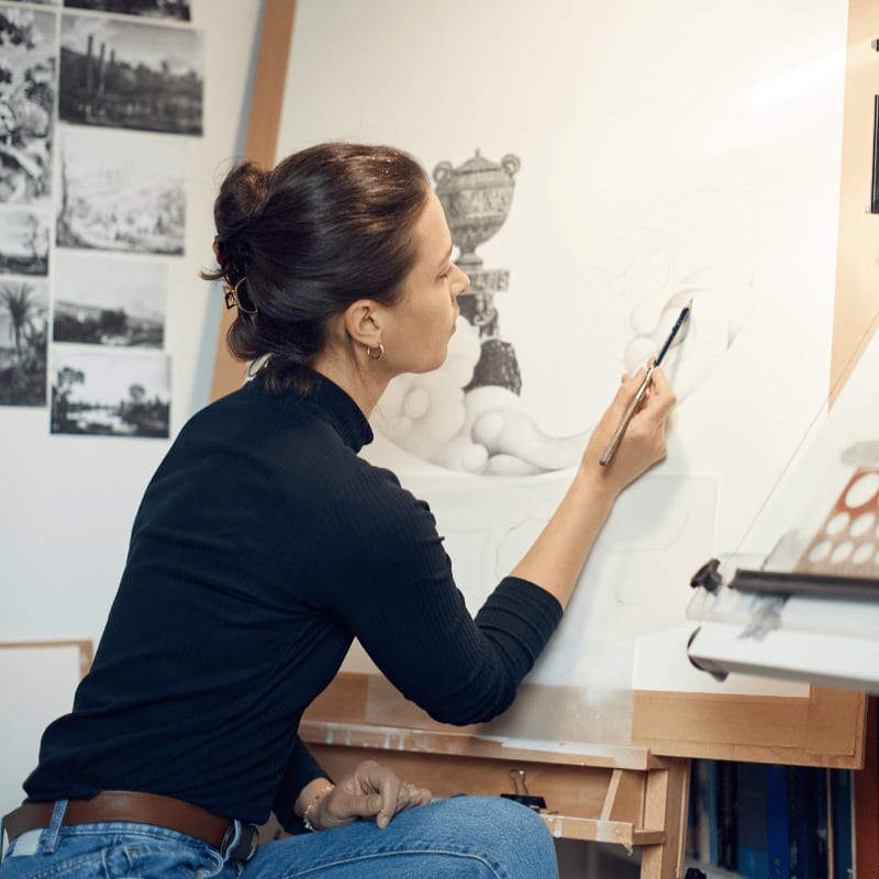 A woman in a black turtleneck and jeans sketches a classical urn on a large canvas, with framed artwork and art supplies visible in the background.