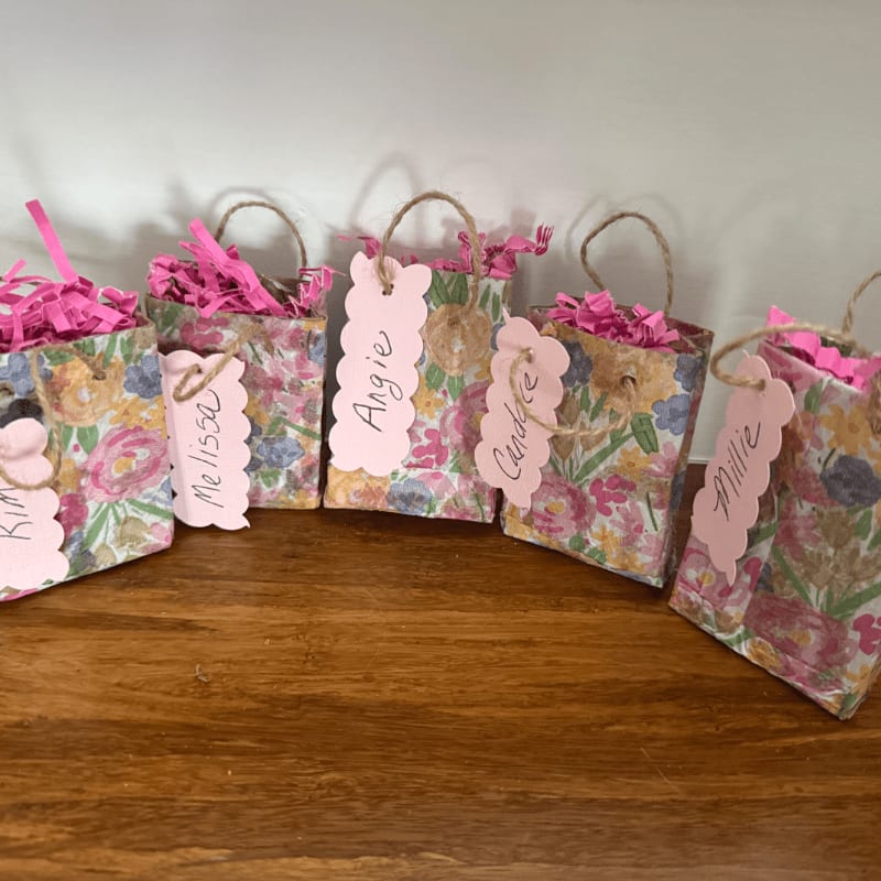 Five small floral gift bags with pink tissue paper and personalized tags sit on a wooden surface in front of a white background.
