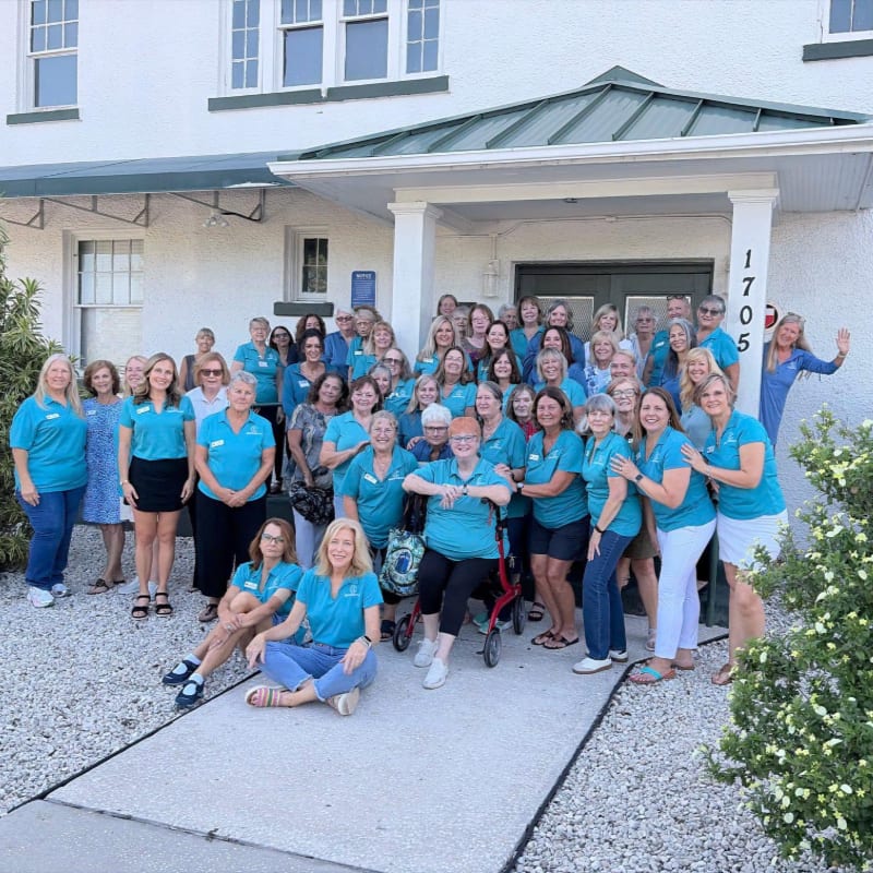 A large group of women in teal shirts pose for a photo on a walkway in front of a white building with a green roof.