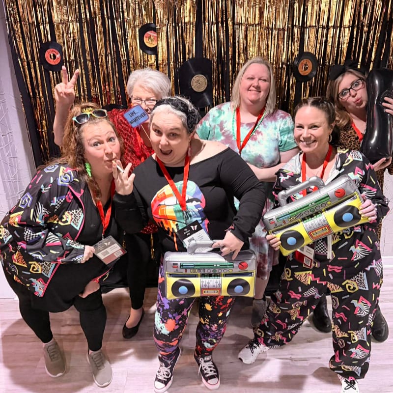 Six women in 1980s-themed outfits pose in front of a gold sequined backdrop adorned with vinyl records.