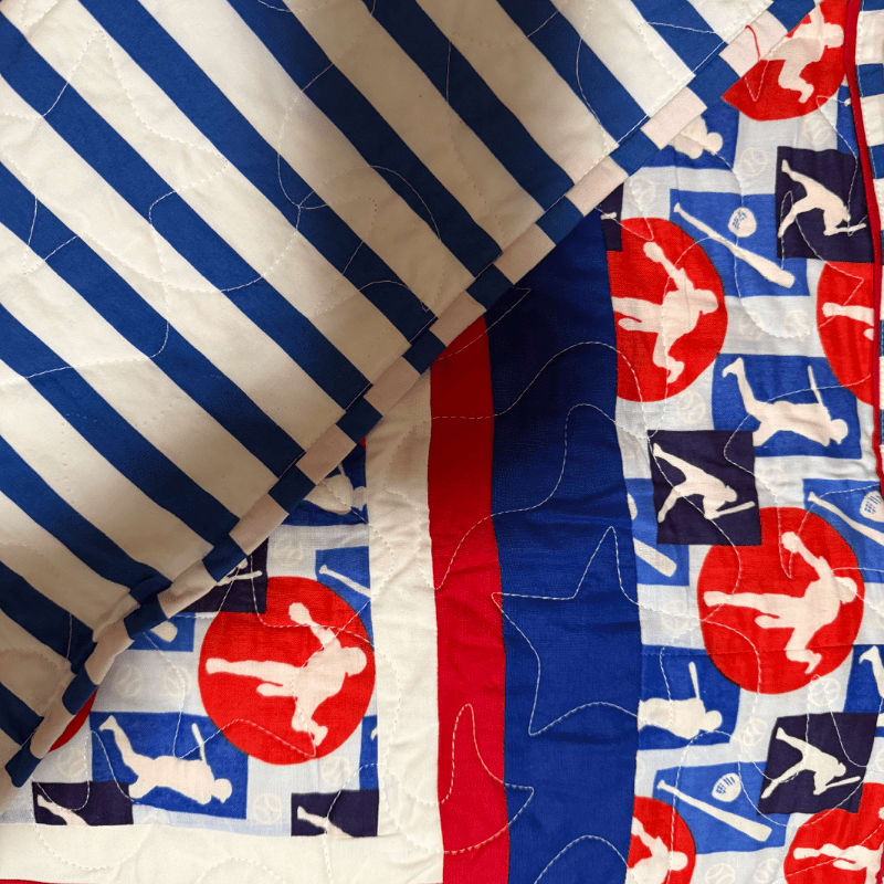 A quilt with a baseball-themed pattern in the background is folded over a striped fabric in the foreground.