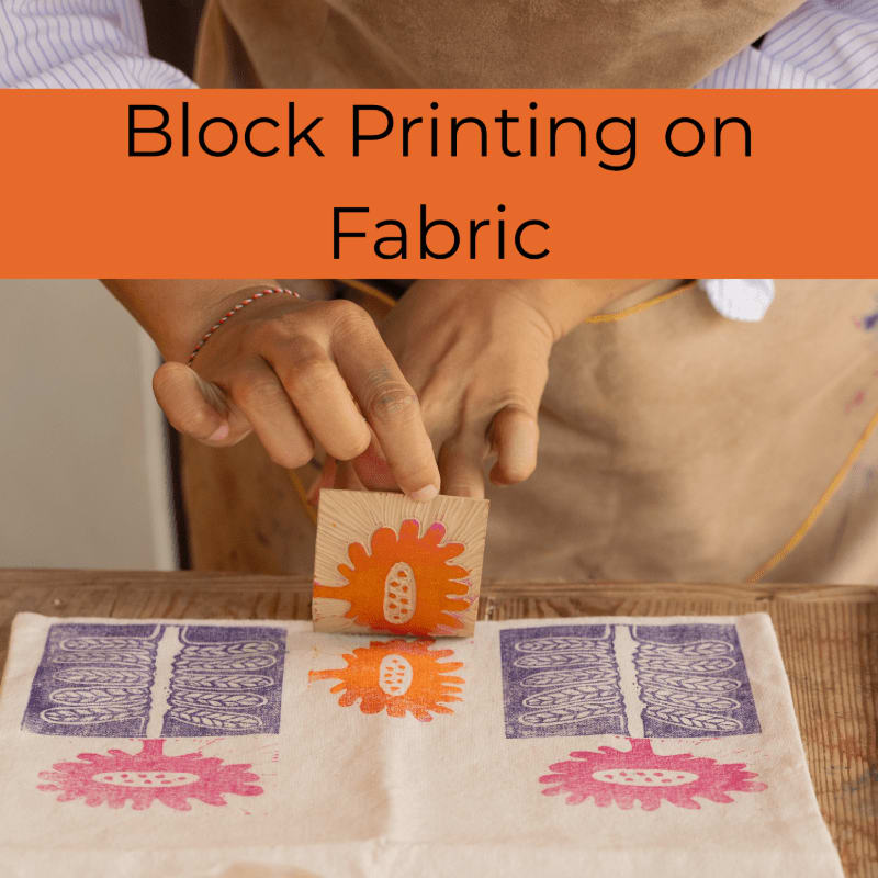 Hands are pressing a carved wooden block with an orange flower design onto a white fabric with purple and pink floral prints, while a brown apron is visible in the background.