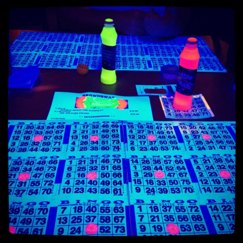 Neon-colored bottles stand on a table covered with bingo cards under blacklight.
