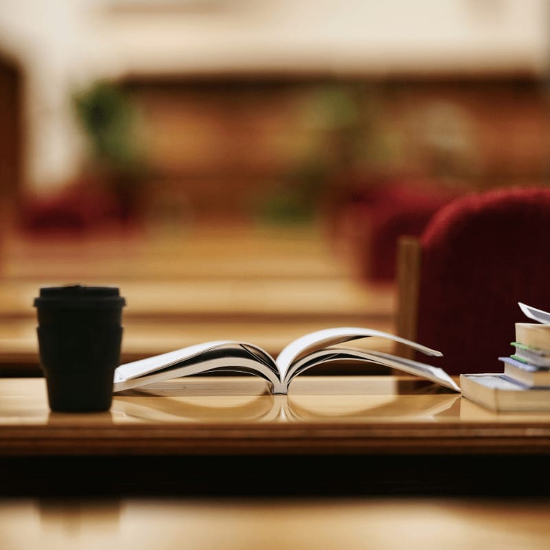 An open book and a coffee cup rest on a wooden table in front of blurred red chairs.