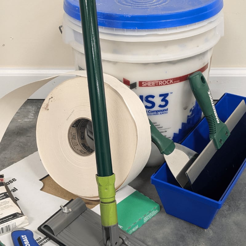 A green-handled drywall taping tool and a roll of tape are in the foreground, with a bucket of joint compound and other tools in the background.