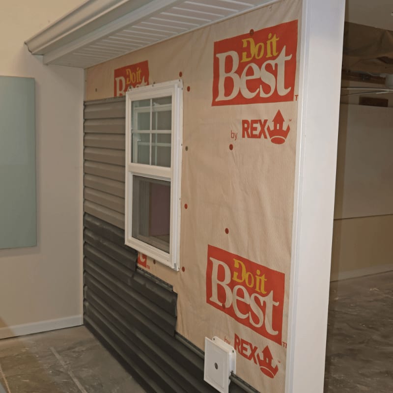 A partially sided wall with a window and house wrap is in the foreground, while a concrete floor and unfinished ceiling are in the background.