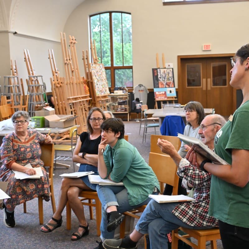 A group of people are gathered in an art studio, with easels and artwork visible in the background.