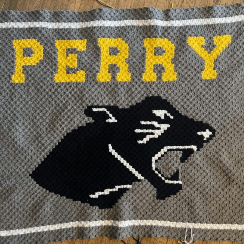 A crocheted blanket with the word "PERRY" in yellow letters above a black panther head on a gray background, bordered by white stripes.