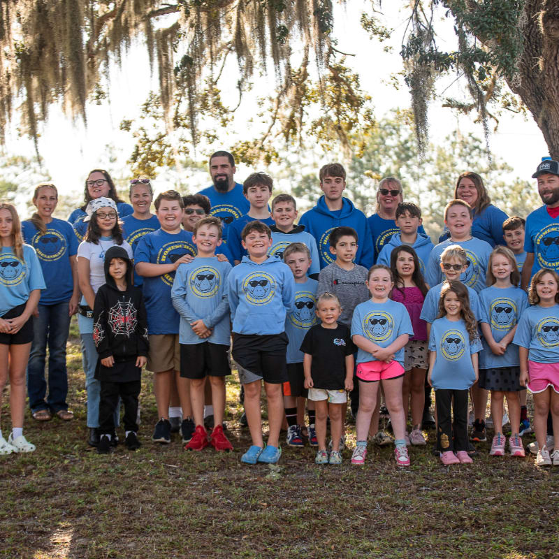 A large group of people, including adults and children, pose for a photo outdoors in front of a large tree with Spanish moss hanging from its branches.