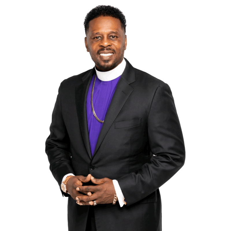 A smiling man in a black suit and purple clerical shirt stands against a plain white background.