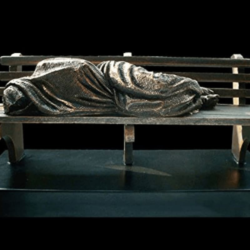 A bronze sculpture of a person sleeping on a park bench is set against a stark black background.
