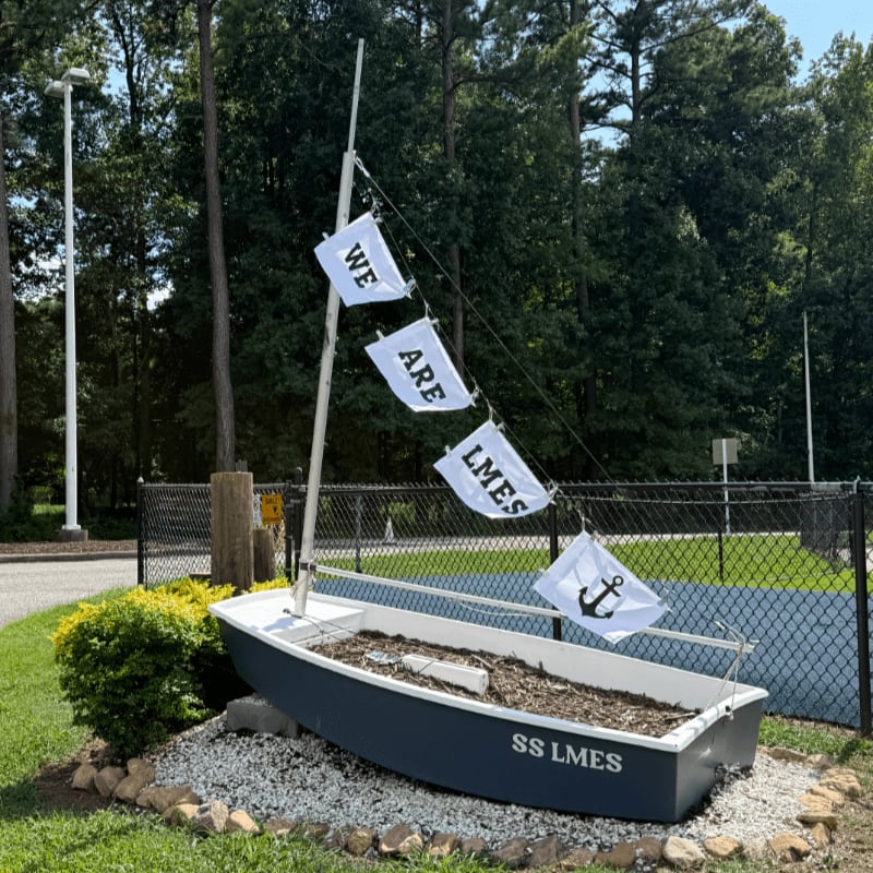 A small, dark blue boat with "SS LMES" painted on its side sits in the foreground, adorned with flags that read "WE ARE LMES" and an anchor, while a lush green forest forms the background.