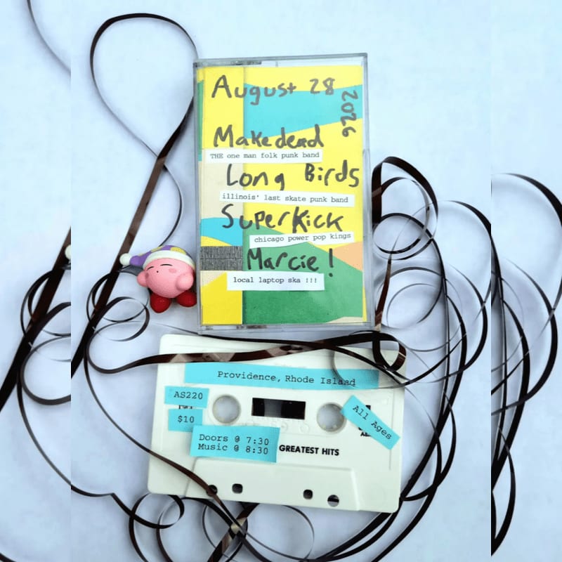 A white cassette tape with handwritten event details sits in the foreground amidst tangled magnetic tape, with a small pink Kirby toy to its left, against a light blue background.
