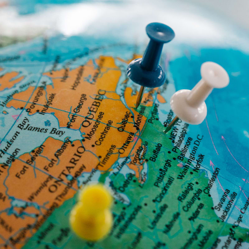 Three pushpins mark locations on a globe, with the foreground showing a blurry yellow pin and the background displaying North America with various cities labeled.