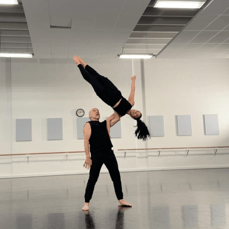 A man in black pants and a sleeveless shirt supports a woman in black athletic wear as she performs an acrobatic dance move, with a dance studio featuring a barre, mirrors, and acoustic panels in the background.