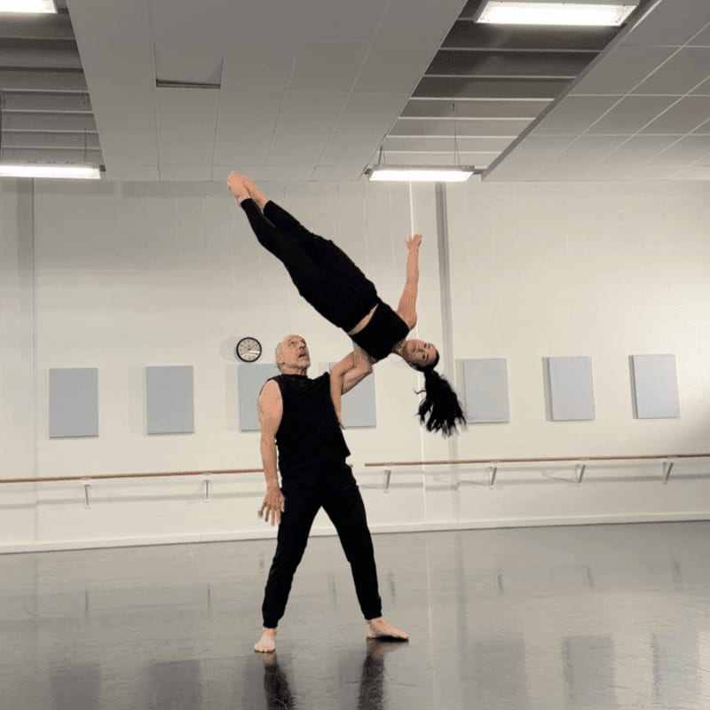 A man in black pants and a sleeveless shirt supports a woman in black athletic wear as she performs an acrobatic dance move, with a dance studio featuring a barre, mirrors, and acoustic panels in the background.
