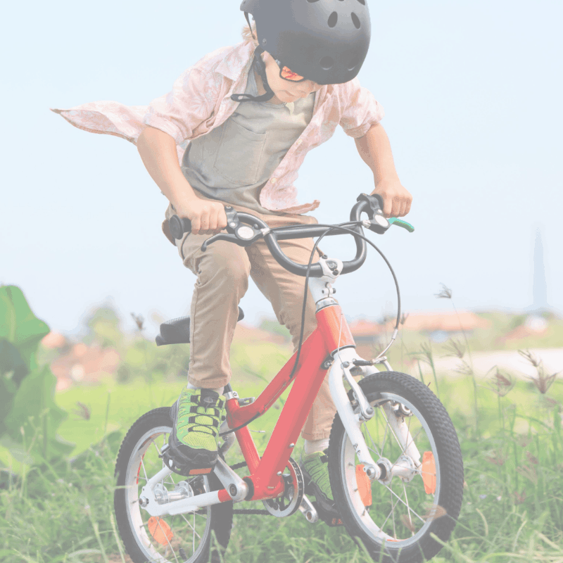 A young boy in a helmet rides a red bicycle through a grassy field with blurred foliage in the background.