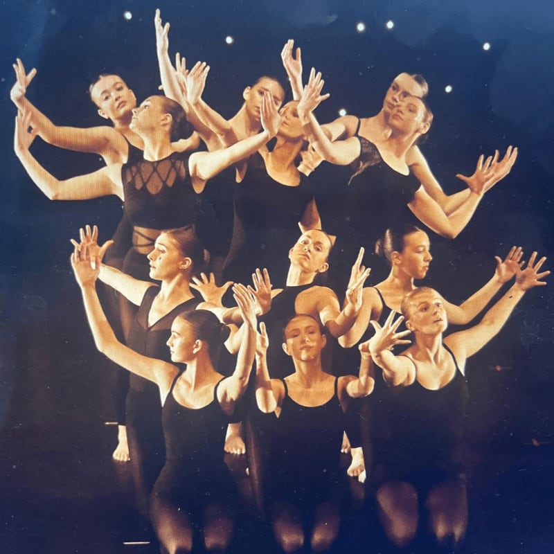 Dancers in black leotards strike poses on a dark stage with a blue spotlight in the background.