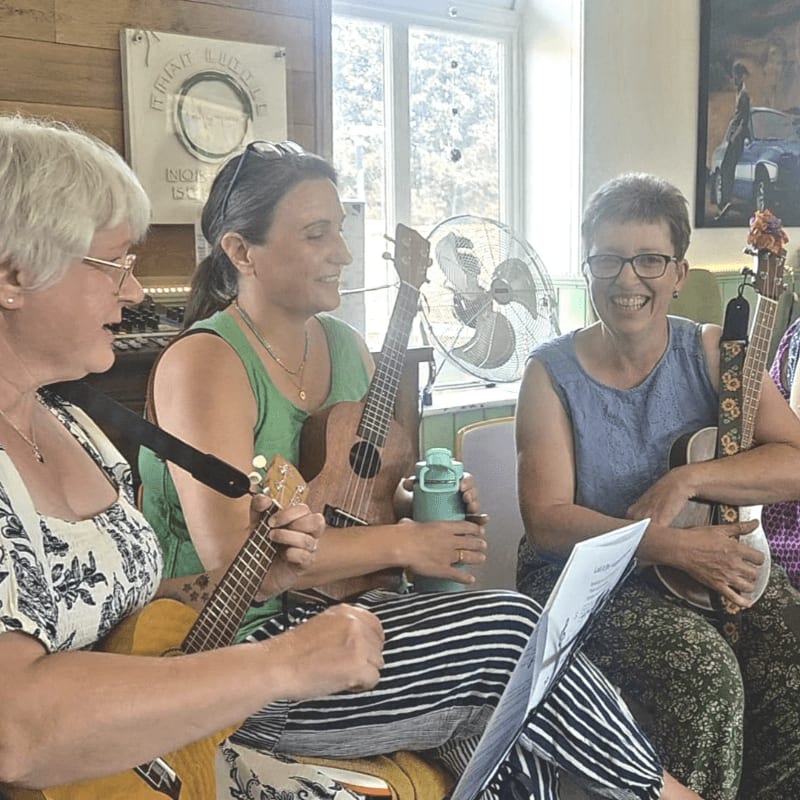Three women are playing ukuleles in a room with a fan and artwork in the background.
