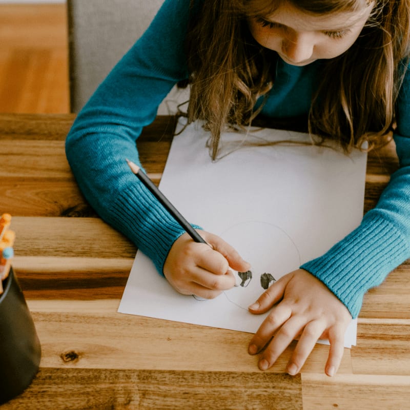 A young girl in a teal sweater draws on a white sheet of paper with a pencil, with a cup of colored pencils to her left and a wooden table beneath.