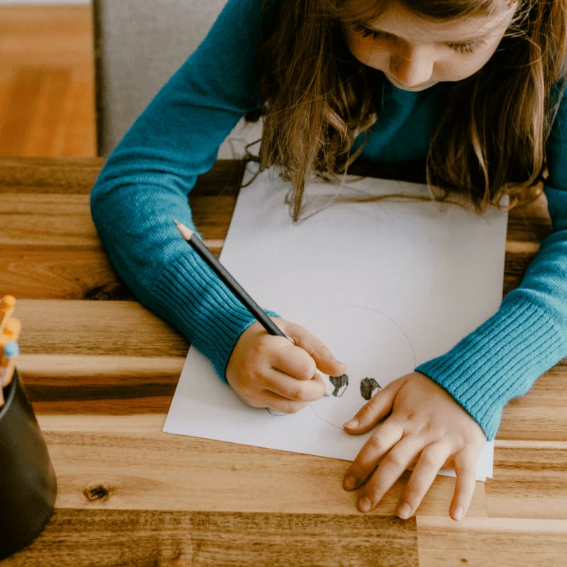 A young girl in a teal sweater draws on a white sheet of paper with a pencil, with a cup of colored pencils to her left and a wooden table beneath.