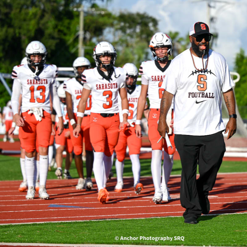 A football coach walks alongside players on a track, with a stadium and trees in the background.