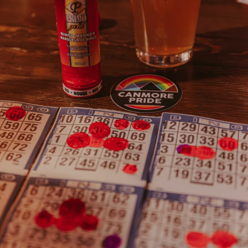 Bingo cards marked with red and purple daubers sit on a wooden table next to a bingo marker and a glass of beer, with a "Canmore Pride" sticker in the background.