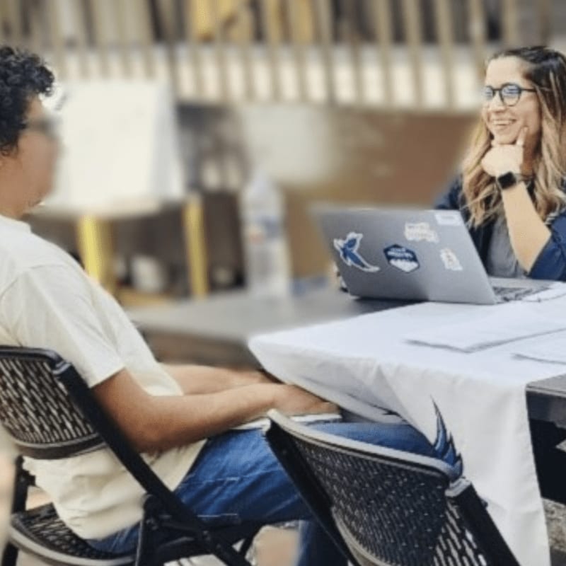 A blurred person in the foreground sits across a table from a smiling woman with glasses, who is looking at a laptop in the background.