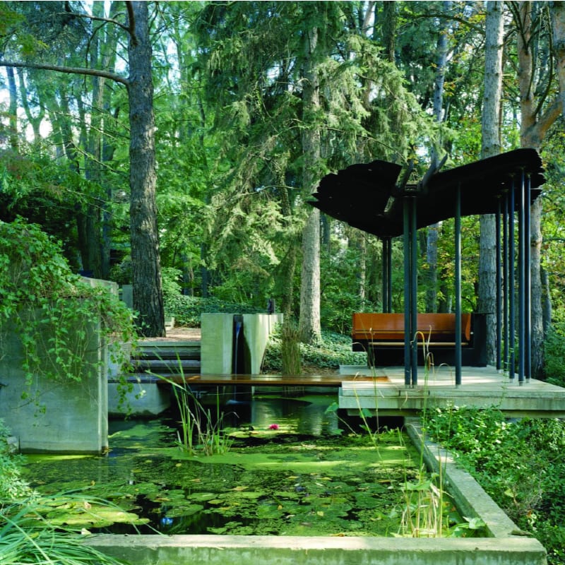 A tranquil pond with lily pads in the foreground is set against a backdrop of a modern open-air pavilion nestled within a lush, green forest.