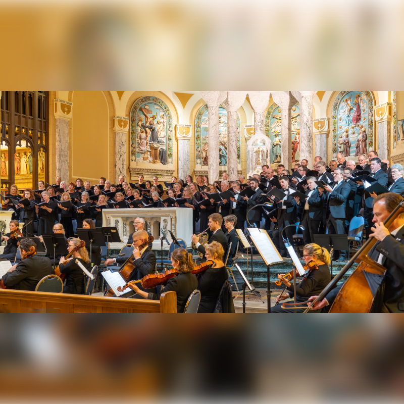 A choir and orchestra perform in a church with ornate altars and paintings in the background.
