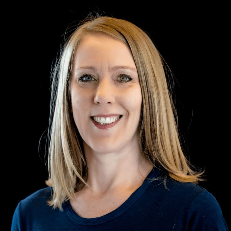 A smiling woman with blonde hair and a dark blue shirt is pictured against a solid black background.