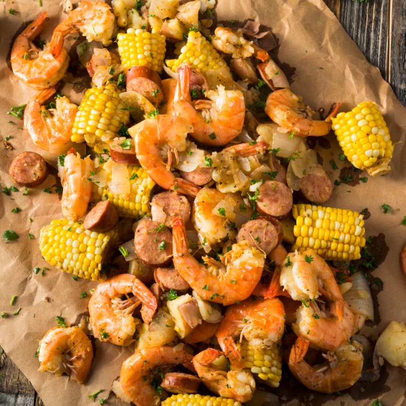 A close-up overhead view of a seafood boil with shrimp, corn, and sausage scattered across brown parchment paper, with a wooden surface visible in the background.
