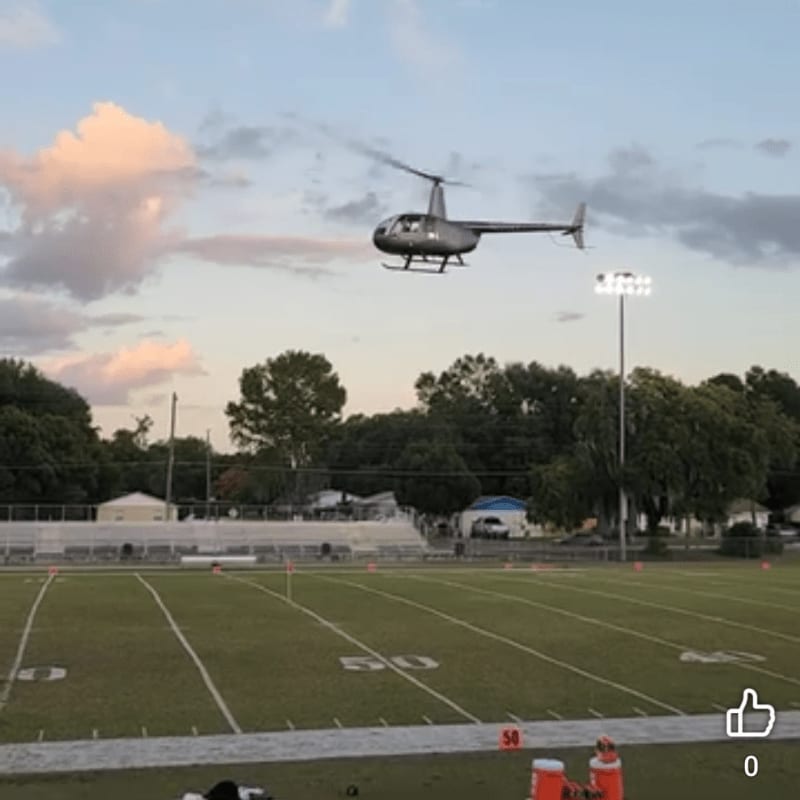 A helicopter flies over a football field with bleachers and trees in the background under a cloudy sky.