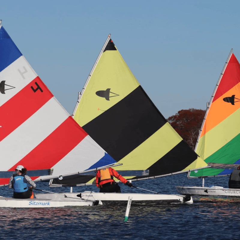 Three colorful sailboats with people in them glide across a calm blue lake with a tree-lined shore in the background.