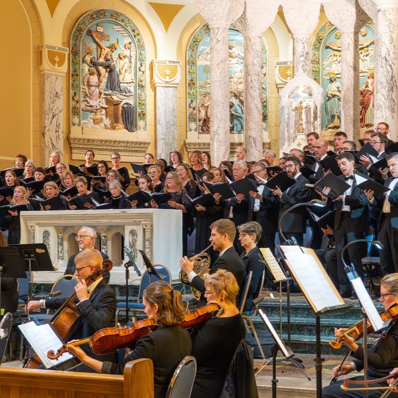 A choir and orchestra perform in a church with ornate arches and religious artwork in the background.